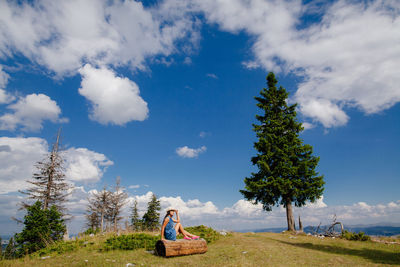 Full length of woman sitting on log over landscape against blue sky