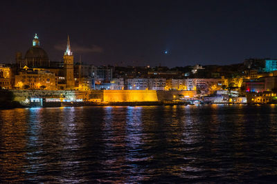 Illuminated cityscape by river against sky at night