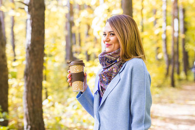Young woman drinking coffee while standing by tree during autumn