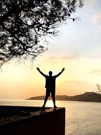 Silhouette man with arms raised standing by sea during sunset