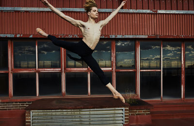 Masterful young blond ballet dancer in black leotard performing exciting jump in splits raising hands up on roof of building