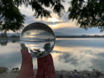 Reflection of person hand holding crystal ball on lake against sky
