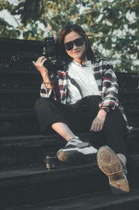 Young woman wearing sunglasses and holding her camera is sitting outdoors 