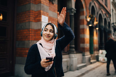 Smiling teenage girl waving hand while listening to headphones on footpath in city