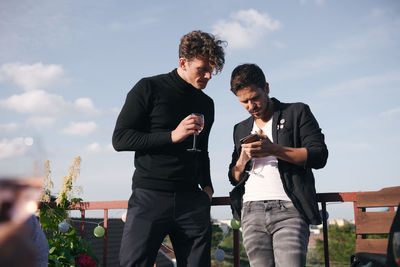 Low angle view of friends using mobile phone while standing against sky on terrace