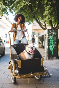 Smiling woman riding bicycle cart with bulldog on cobbled street