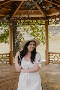 Portrait of beautiful young woman wearing white dress standing under wooden pavilion in park.