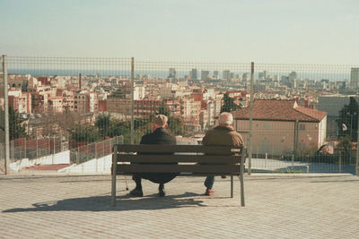 Rear view of men sitting on bench in city against clear sky