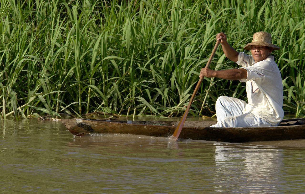 water, one person, nature, hat, full length, adult, plant, holding, sitting, day, lake, clothing, waterfront, reflection, nautical vessel, side view, occupation, casual clothing, outdoors, farmer