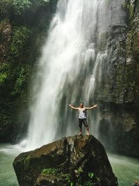 Young man with arms outstretched standing on rock against waterfall