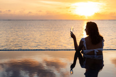 Attractive woman on a infinity pool near the ocean with a glass of champagne.back view