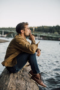 Side view of man smoking while sitting on log by river against clear sky