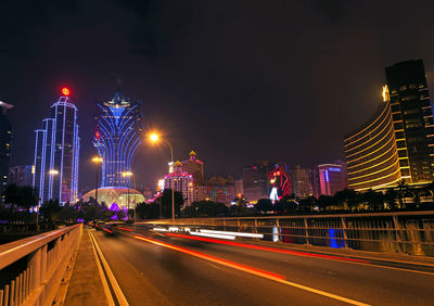 Light trails on road by illuminated buildings against sky at night