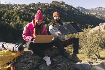 Young woman using laptop sitting by man adjusting solar panel on sunny day