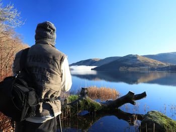 Rear view of man standing by lake against clear blue sky