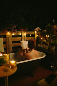 Back view of unrecognizable naked female covering face with book while sitting in bath placed in patio with glowing garland in evening time