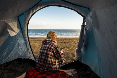 Woman sitting in tent at beach against sky