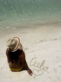 High angle view of sensuous woman wearing hat while sitting on sand at beach