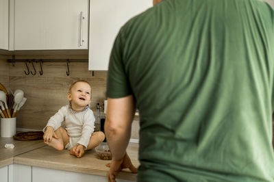 Happy cute baby girl sitting on counter looking at father standing in kitchen