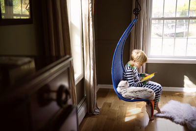 Side view of boy reading book while sitting on swing in bedroom