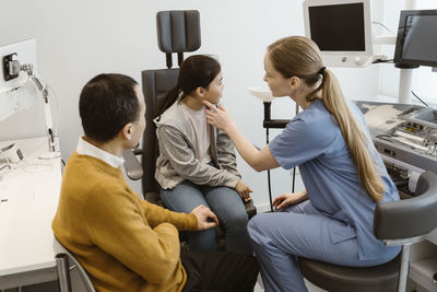 Side view of female pediatrician examining girl sitting on chair by father in medical clinic