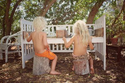 Rear view of shirtless children playing while sitting on tree stumps in yard