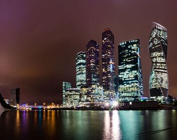 Illuminated modern buildings by river against sky at night