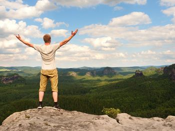 Happy man with open rised arms. satisfy hiker in grey shirt and trekking shorts on cliff watch down