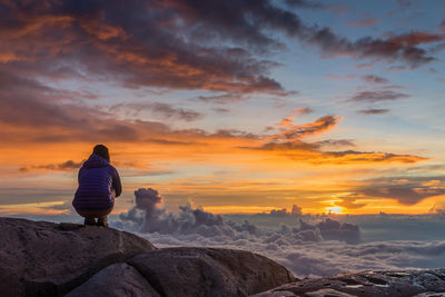 Rear view of woman sitting on rock against sky during sunset