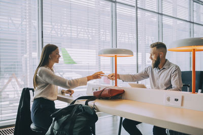 Smiling businessman giving charger to young businesswoman while sitting at airport