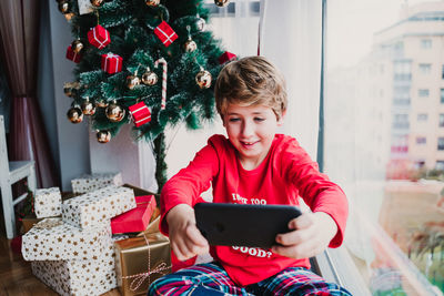 Portrait of beautiful kid boy at home by the christmas tree and using mobile phone