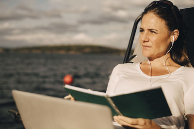 Thoughtful woman with laptop holding diary while looking away against lake