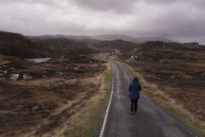 Full length rear view of person walking on empty road against cloudy sky