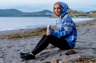 Young woman holding insulated drink container while sitting at beach