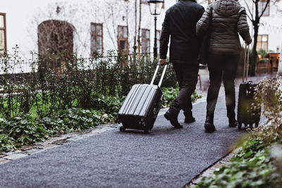 Rear view of senior couple walking on footpath with luggage outside hotel