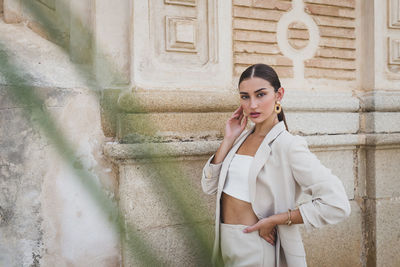 Confident young ethnic female in fashionable beige suit and white top standing near stone wall of urban building and looking at camera