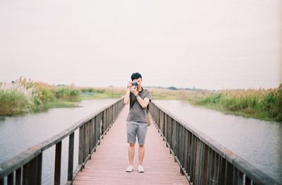 Full length of photographer photographing on footbridge over river against clear sky