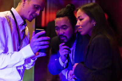 Businessman sharing smart phone with male and female colleagues in illuminated convention center