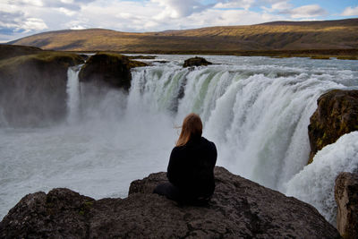 Rear view of woman sitting on rock against godafoss falls