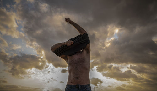 Low angle view of man taking off cloth against cloudy sky