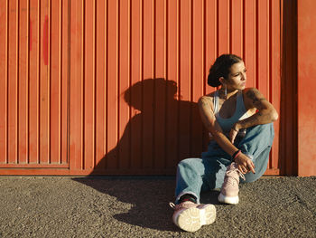 Confident young female with tattoos wearing stylish outfit sitting on floor near red wall and looking away