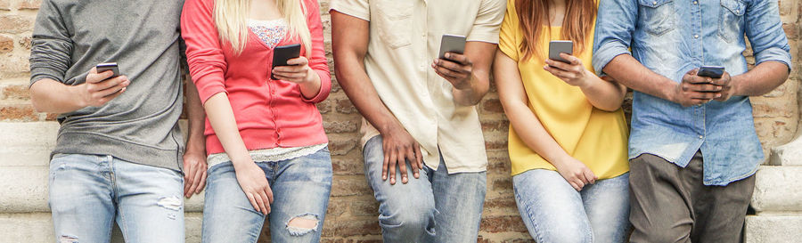 Midsection of friends using smart phones while standing against wall