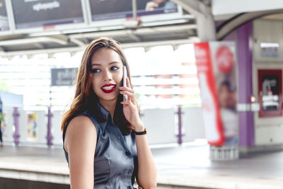 Smiling young woman using mobile phone while standing at railroad station 