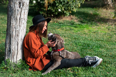 Woman sitting by tree trunk with pet dog in park