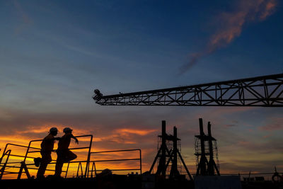 Workers leaning on railing by silhouette crane at construction site against sky during sunset