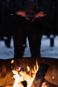 Crop anonymous hiker holding hands over burning campfire while warming up during camping in evening time in winter forest