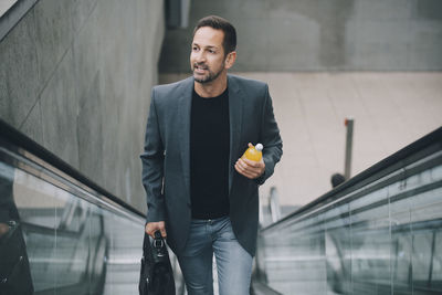 Confident businessman with drink looking away while moving on escalator in subway