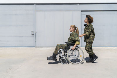 Young female army soldier pushing colleague on wheelchair at military base during sunny day