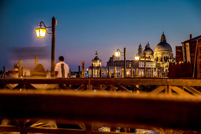 Illuminated buildings by river against sky at dusk