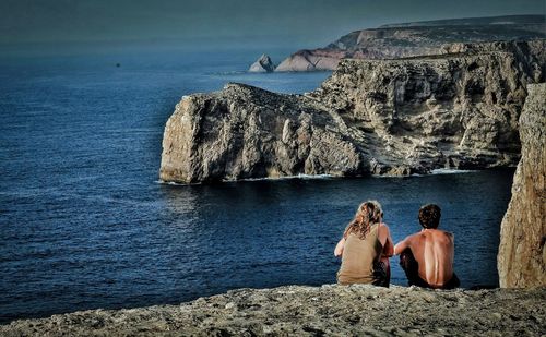 Rear view of couple sitting on cliff by sea